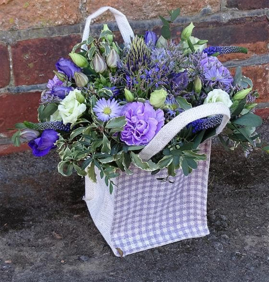 Catherine Wheel Floral Bag Arrangement. This purple and lilac arrangement is made up of Lisianthus, Veronica, September Flower, Carnations and seasonal flowers and foliage.  The flowers are presented in a checked hessian bag. This makes an excellent Autumn gift for a new Mum, someone who is ill or someone who has just moved into their new home who needs a ready made arrangement are in floral foam which is easy to care for.