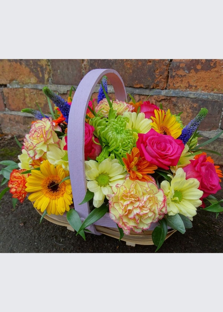 Catherine Cookson Flower Basket. A fabulous vibrant trug basket, filled with bright purple, lime green, yellow, orange and cerise seasonal flowers including Roses, Gerberas, Carnations and Chrysanthemums with seasonal flowers and foliages. This makes an ideal Spring gift for any occasion as it is a ready made arrangement in floral foam which is easy to care for.