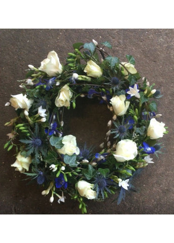 Countryside Walk Funeral Wreath by Make Their Day florist. This detailed modern, textured funeral wreath is filled with blue and white Roses, Freesia, Eryngium, Delphinium, lavender and seasonal twigs and foliages. This loose design showcases this beautiful medley of a Cotswold countryside walk in colours and textures.