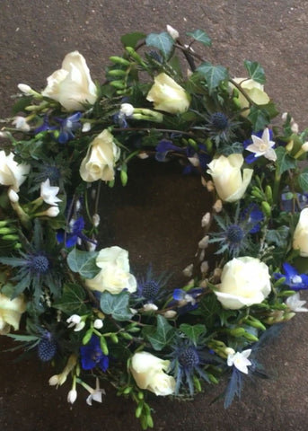 Countryside Walk Funeral Wreath by Make Their Day florist. This detailed modern, textured funeral wreath is filled with blue and white Roses, Freesia, Eryngium, Delphinium, lavender and seasonal twigs and foliages. This loose design showcases this beautiful medley of a Cotswold countryside walk in colours and textures.