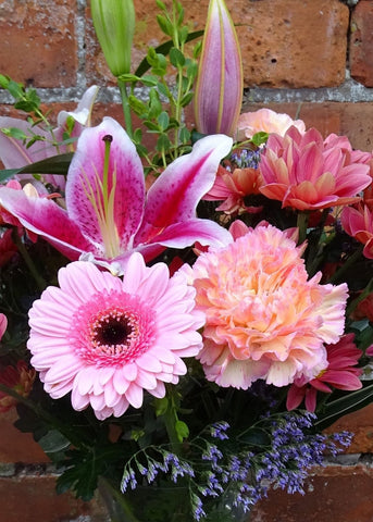Shukran Bouquet in a Vase. This hourglass shaped glass vase is filled with pink flowers with hint of purple arranged in a vintage style. It includes a pink Lily, Gerberas, and Chrysanthemums with other seasonal flowers and foliages. This makes for a gorgeous birthday gift.