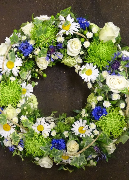 Summer Meadow Funeral Wreath: This loose wreath funeral tribute of green, white, and blue flowers is filled with green Chrysanthemum blooms, Carnations and foliage, white Roses, Lisianthus, Leucanthemums, Veronicas and Chrysanthemums and blue Cornflowers and filled in with delicate seasonal berries, twigs, flowers and foliage.