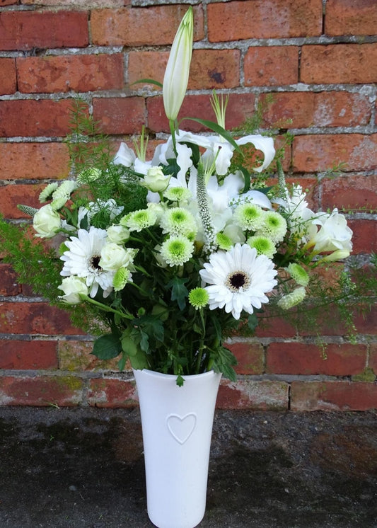 Virginia Woolf Bouquet in a Vase. This white slim waisted glass vase is filled to bursting with white flowers arranged in a vintage style. It includes White Lilies, and Gerberas, Cream Spray Roses and Green Chrysanthemums, seasonal flowers and complemented with delicate foliages combined to create a unique gift.