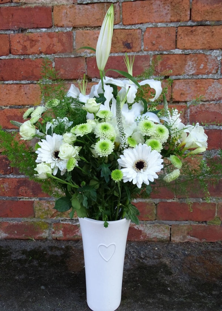 Virginia Woolf Bouquet in a Vase. This white slim waisted glass vase is filled to bursting with white flowers arranged in a vintage style. It includes White Lilies, and Gerberas, Cream Spray Roses and Green Chrysanthemums, seasonal flowers and complemented with delicate foliages combined to create a unique gift.